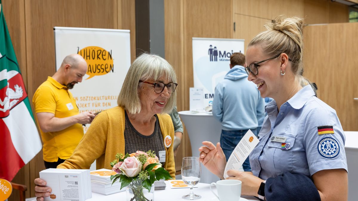 Projekt "Zuhören.Draußen" © Jörg Farys / BBE Zwei Frauen, eine ältere und eine jüngere, unterhalten sich an einem Stehtisch. Im Hintergrund sind Rollups aufgestellt.