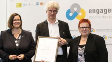Götz Peter Kaiser bei der Erennung zum Engagement-Botschafter 2016 Zwei Frauen und ein Mann stehen vor einer Logowand. Der Mann in der Mitte hält eine Urkunde vor sich.