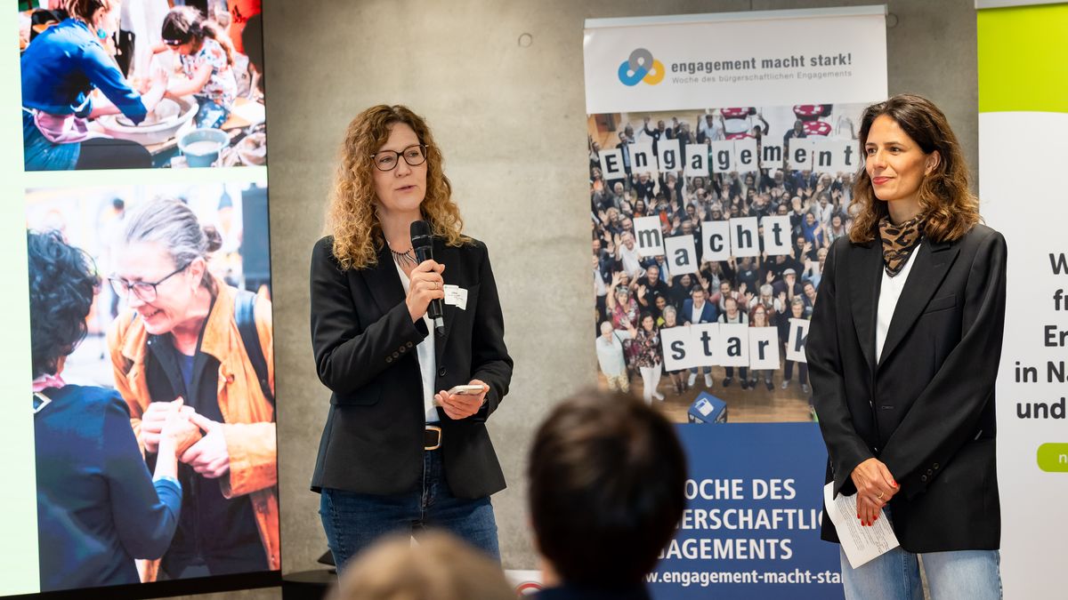 Begrüßung durch BBE und nebenan.de Stiftung © Jörg Farys Zwei Frauen stehen vor Publikum mit Mikrofon. Hinter ihnen stehen zwei Rollups.