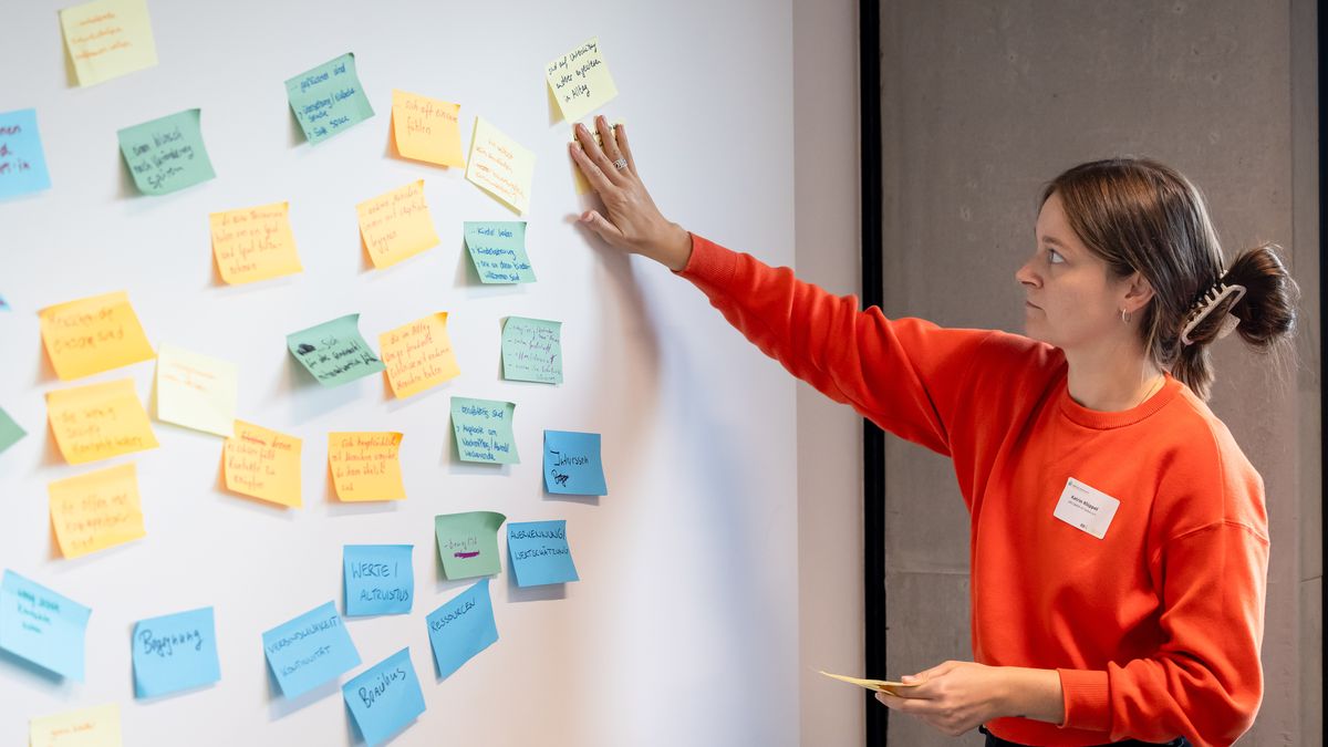 Workshopsituation © Jörg Farys Eine Frau steht an einer Wand mit beschriebenen Haftnotizen und klebt weitere Zettel an die Wand.