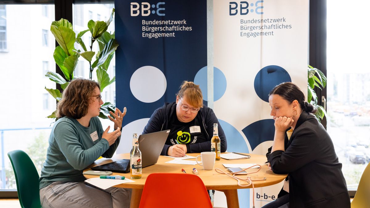 Workshopsituation Gruppenarbeit © Jörg Farys Drei Frauen sitzen an einem Tisch und erarbeiten etwas zu einer Aufgabe. Im Hintergrund stehen zwei Rollups des BBE.