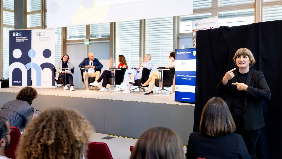 Podiumsdiskussion mit DGS © Jörg Farys / BBE Fünf Menschen sitzen auf einer Bühne. Im Vordergrund ist ein wenig vom Publikum zu sehen. Rechts steht eine Frau und übersetzt in Deutsche Gebärdensprache.