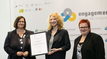 Heike-Melba Fendel bei der Ernennung zur Engagement-Botschafterin 2016 Drei Frauen vor einer Logowand. Die Frau in der Mitte hält eine Urkunde vor sich.