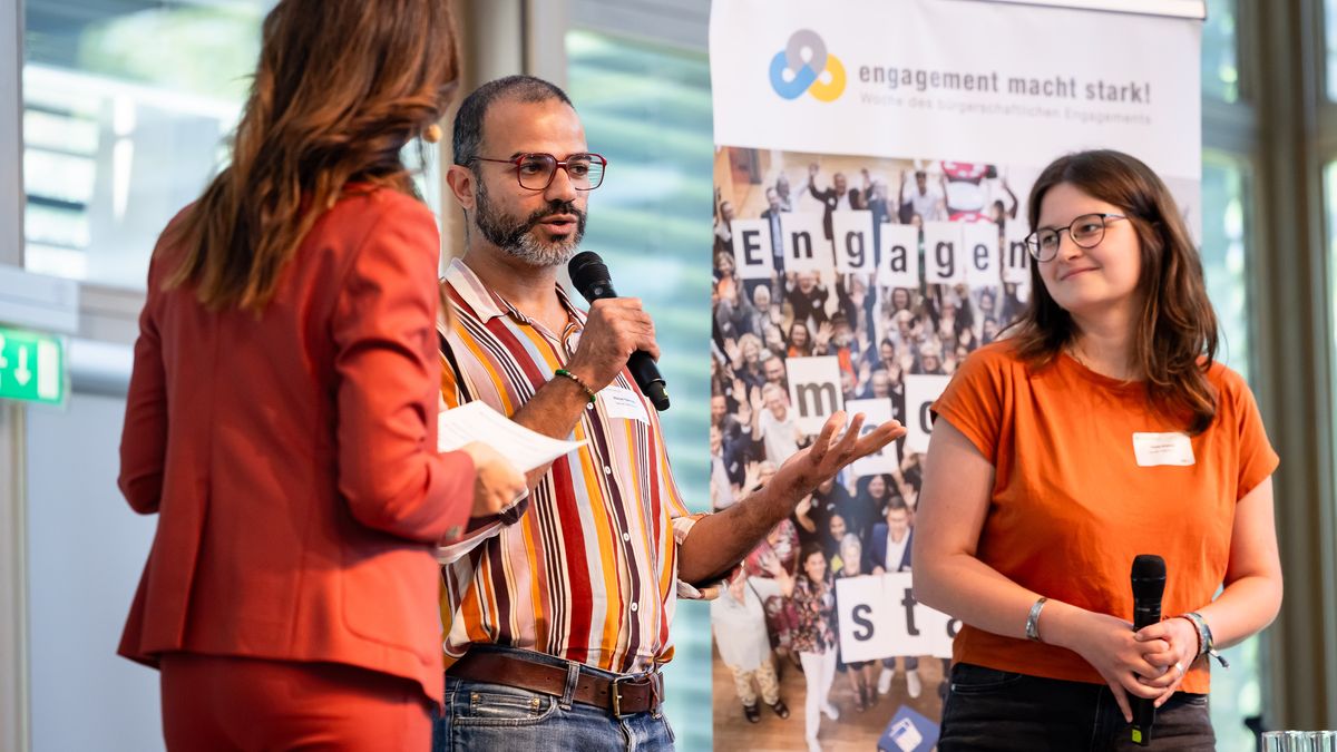 Über den Tellerrand e. V. © Jörg Farys / BBE Drei Personen stehen auf einer Bühne und unterhalten sich. Der Mann in der Mitte hält ein Mikro und spricht.