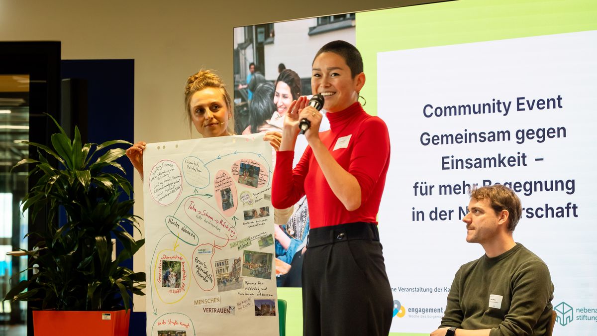 Gemeinsamer Abschluss © Jörg Farys Eine Frau steht vor einem großen Bildschirm und spricht in ein Mikrofon. Eine Frau neben ihr hält ein Poster.