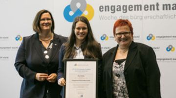 Maxi Kluttig bei der Ernennung zur Engagement-Botschafterin 2016 Drei Frauen stehen vor einer Logowand. Die Frau in der Mitte hält eine Urkunde vor sich.