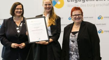 Tjorven Reisener bei der Ernennung zur Engagement-Botschafterin 2016 Drei Frauen stehen vor einer Logowand. Die Frau in der Mitte hält eine Urkunde vor sich.
